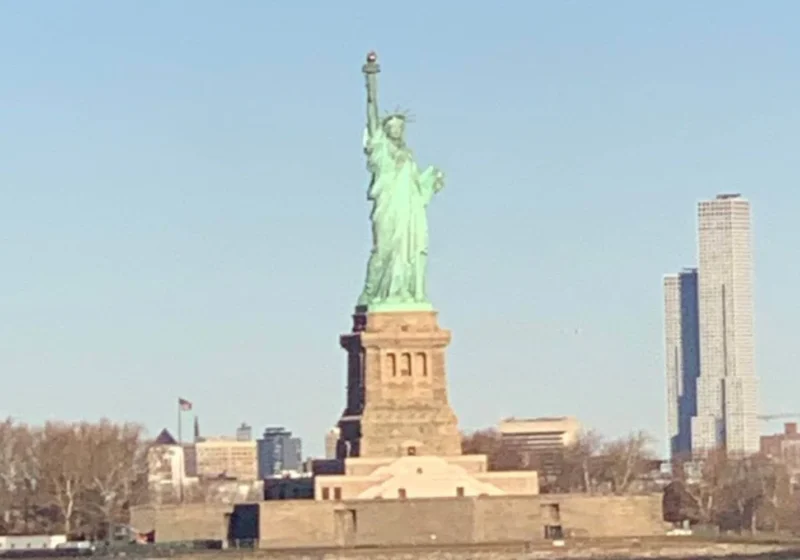 Distant view of the Statue of Liberty standing tall on Liberty Island with a clear blue sky behind it and modern skyscrapers visible in the background. The statue's green patina contrasts with the stone pedestal and surrounding bare trees.