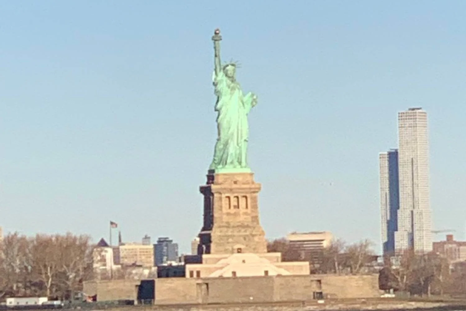 Distant view of the Statue of Liberty standing tall on Liberty Island with a clear blue sky behind it and modern skyscrapers visible in the background. The statue's green patina contrasts with the stone pedestal and surrounding bare trees.