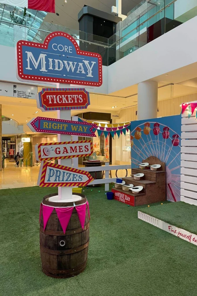 Colorful directional sign inside a Calgary mall reads “CORE Midway,” pointing to “Tickets,” “Games,” “Prizes,” and more, set beside carnival decor like barrels and a small Ferris wheel mural.