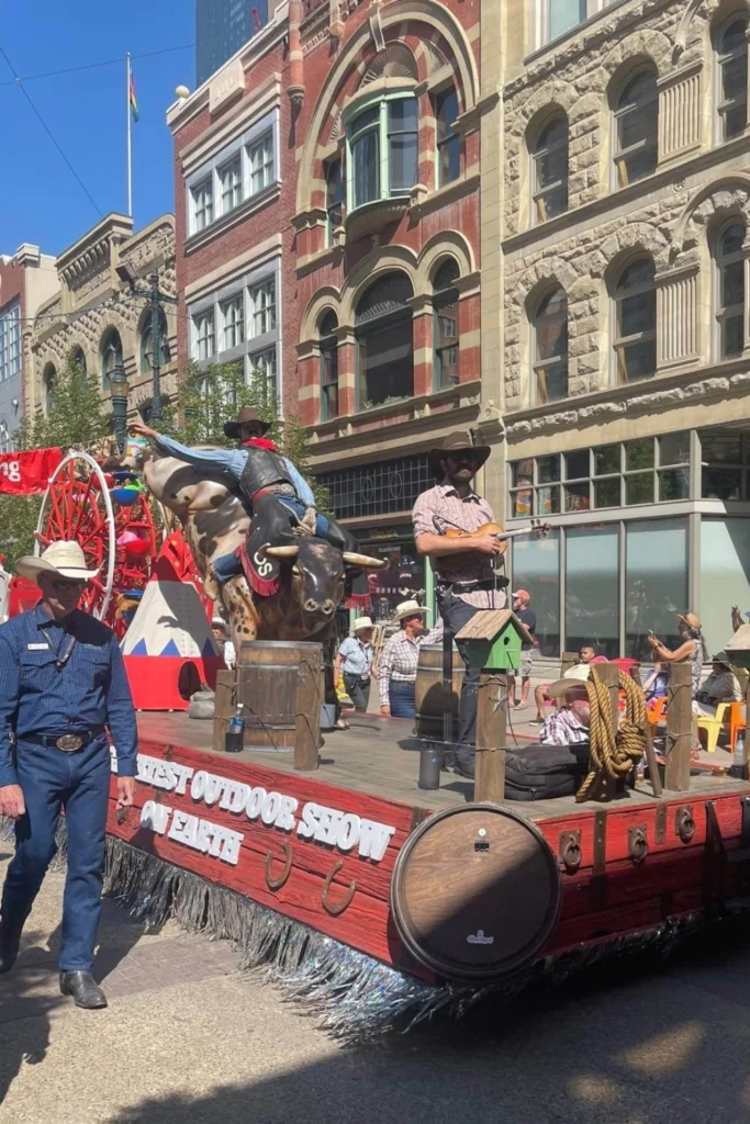 A parade float themed "The Greatest Outdoor Show on Earth" features a life-size model of a cowboy riding a bucking bull, with musicians and props on board. Historic stone and brick buildings line the street as spectators watch under a clear blue sky.