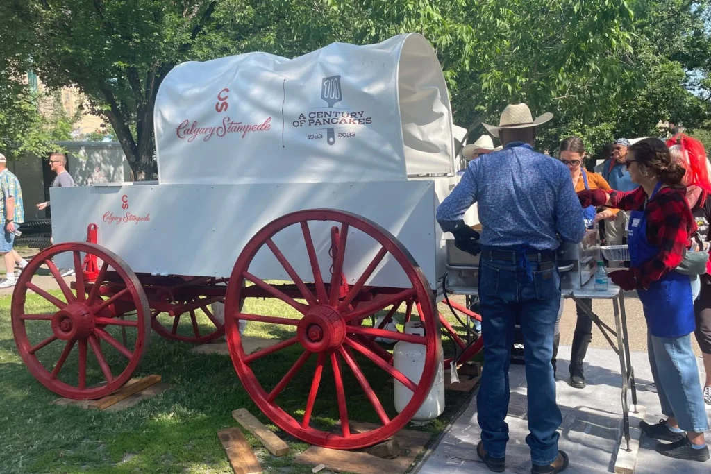A white Calgary Stampede chuckwagon with bright red wheels and "A Century of Pancakes 1923–2023" printed on its cover is surrounded by volunteers cooking pancakes on griddles. People in cowboy hats and aprons prepare and serve breakfast outdoors.