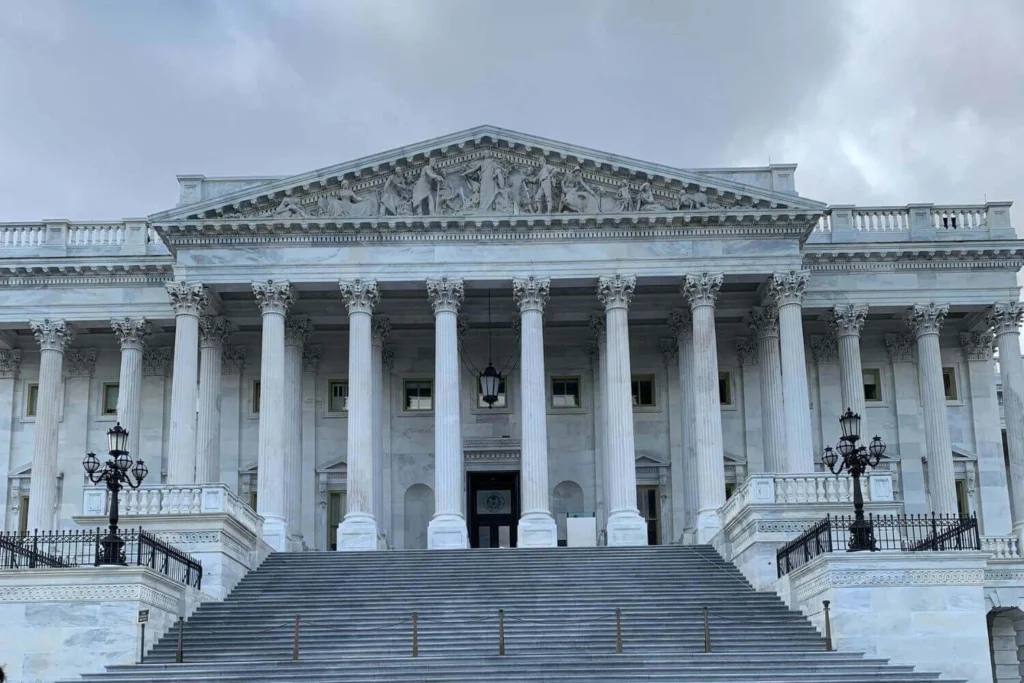 Straight-on view of the U.S. Capitol East Front entrance in Washington D.C., with neoclassical columns and dramatic gray clouds overhead, symbolizing institutional power and government control in 2025 America.
