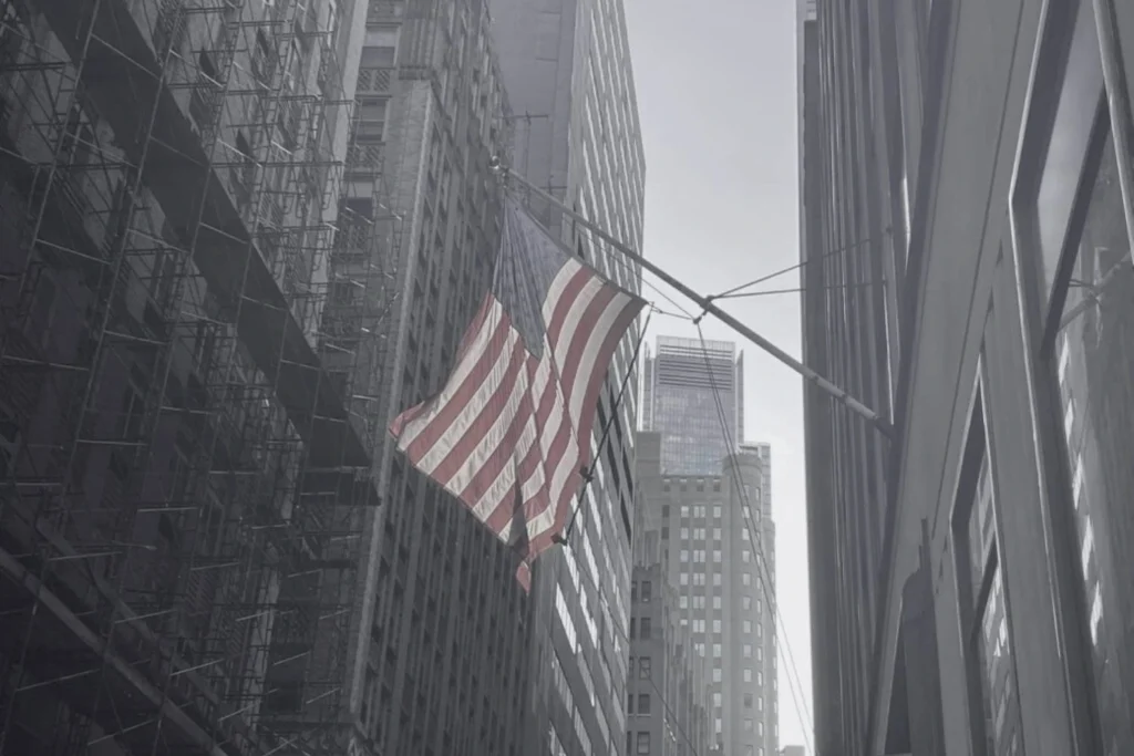 Grey scaled photo of an American flag that waves prominently between tall urban buildings in downtown New York City symbolising patriotism.