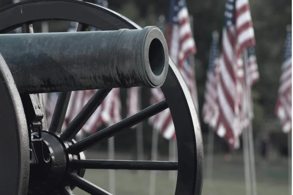 Grey scaled close-up of an old bronze cannon wheel and barrel pointing slightly upward, with a field of blurred American flags waving in the background to symbolize far-right desires to return to the days of the confederacy.