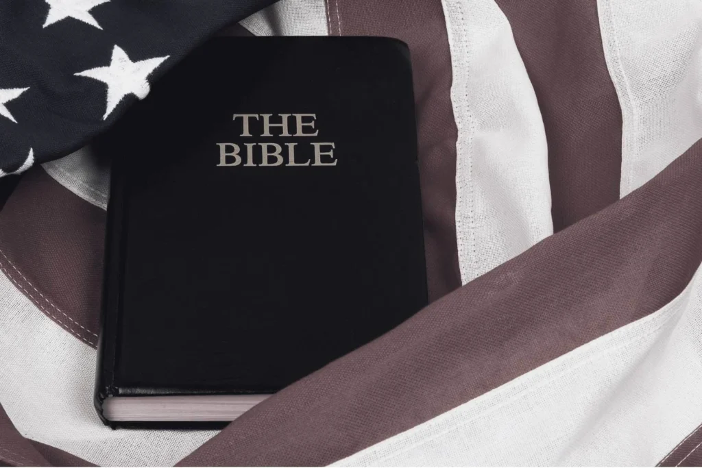 Darkened image of a black-covered Bible with gold lettering lying atop a crumpled American flag, visually representing the close alignment of Christianity and national identity central to Christian nationalism in America.