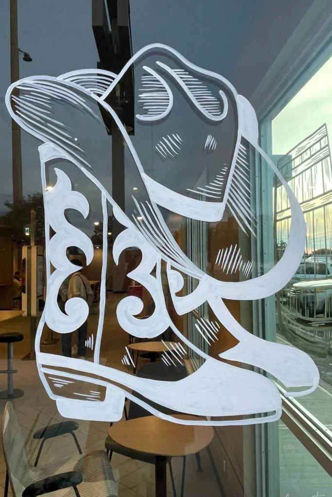 Hand-drawn white outline of ornate cowboy boots and a wide-brimmed hat on a restaurant window, with reflections of tables and city street in Calgary for Stampede.