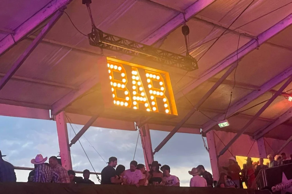 Neon-lit BAR sign inside Cowboys Music Festival tent with festival-goers hanging out at the bar area.
