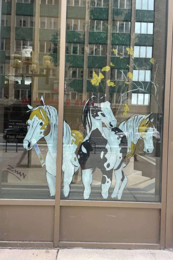 Three painted horses with detailed spots and manes, set against a reflective downtown window in Calgary with falling yellow leaves.