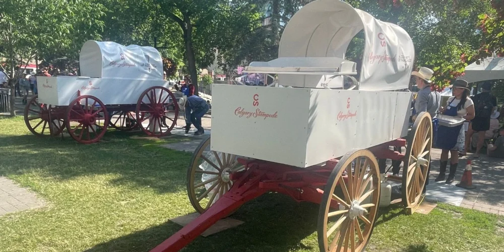 Two white Calgary Stampede chuckwagons sit on a grassy area, one closer to the foreground with "Calgary Stampede" and "A Century of Pancakes 1923–2023" logos visible. Volunteers and attendees in western wear gather nearby under sunny skies.