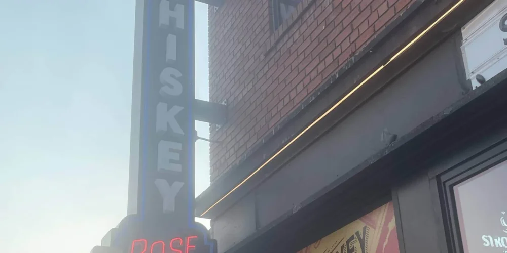 A street view of the Whiskey Rose Saloon featuring a bold neon sign with the words “WHISKEY ROSE SALOON” glowing against a brick building façade.