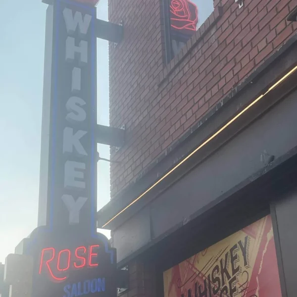 A street view of the Whiskey Rose Saloon featuring a bold neon sign with the words “WHISKEY ROSE SALOON” glowing against a brick building façade.