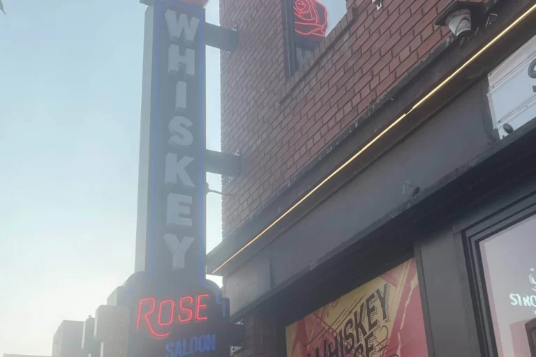 A street view of the Whiskey Rose Saloon featuring a bold neon sign with the words &ldquo;WHISKEY ROSE SALOON&rdquo; glowing against a brick building fa&ccedil;ade.