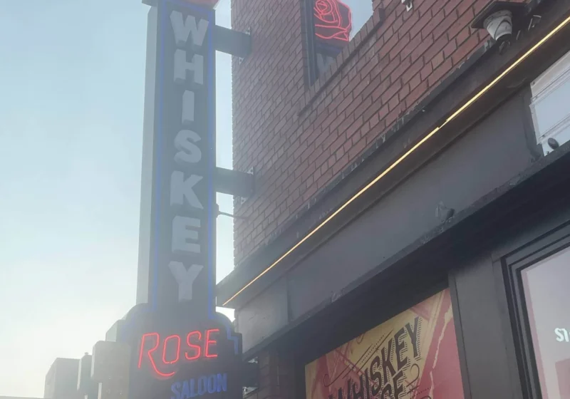 A street view of the Whiskey Rose Saloon featuring a bold neon sign with the words “WHISKEY ROSE SALOON” glowing against a brick building façade.