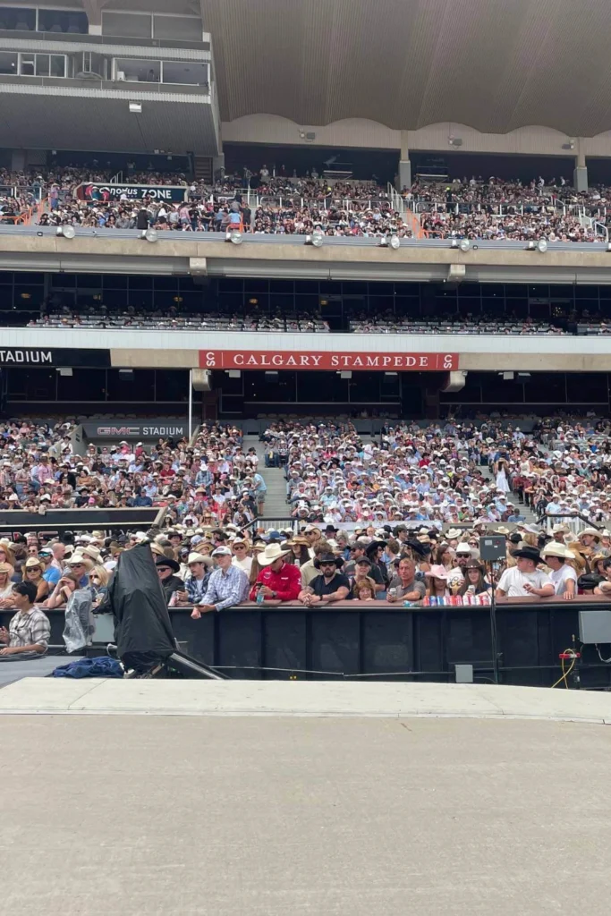 A massive crowd fills the GMC Stadium grandstand at the Calgary Stampede, with spectators wearing cowboy hats and western attire, all facing the stage or arena watching the rodeo.