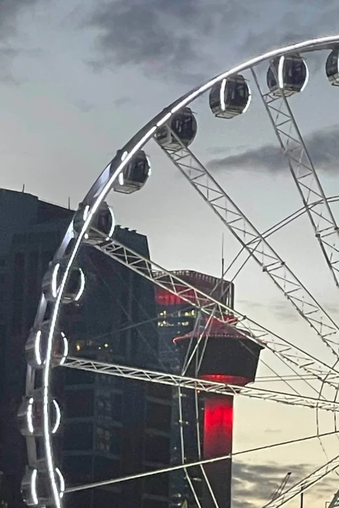 Close-up of the illuminated Calgary Stampede Ferris wheel at dusk, with the Calgary Tower glowing red in the background against a moody evening sky.