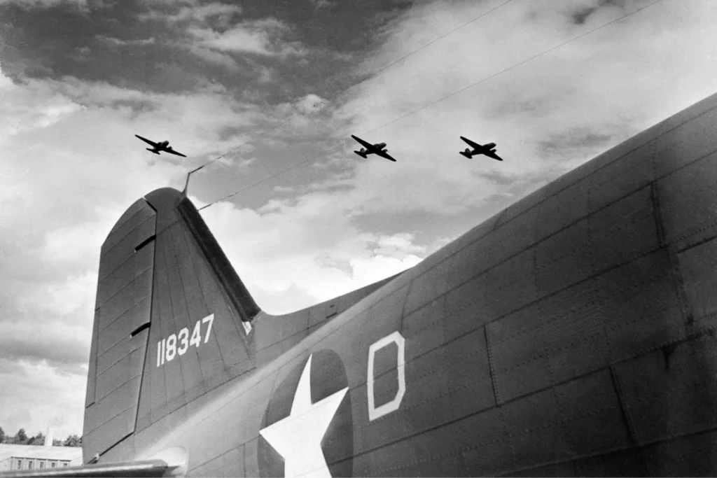 Black-and-white photo of three U.S. military planes flying in formation over England during World War II, seen from behind the tail of a larger aircraft with the U.S. Air Force insignia visible on its fuselage.