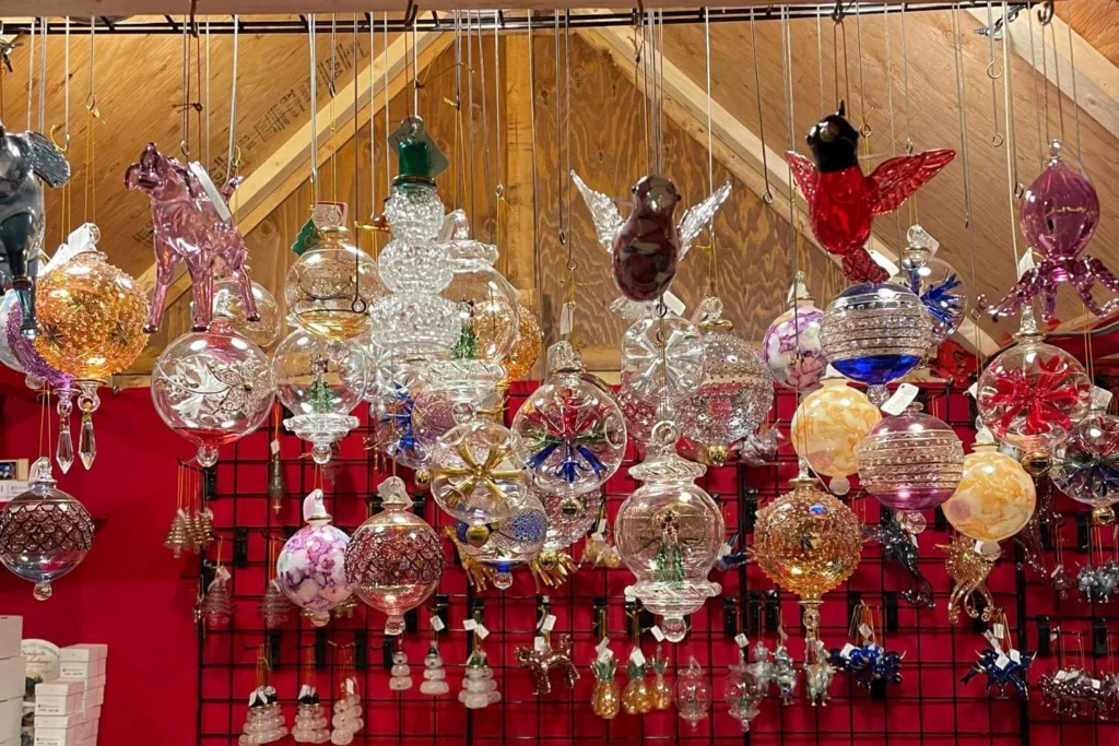 Colourful display of handcrafted glass Christmas ornaments hanging in rows at a market stall at Vancouver Christmas Market, featuring baubles, animals, snowmen, birds, and intricate shapes under warm wooden rafters.