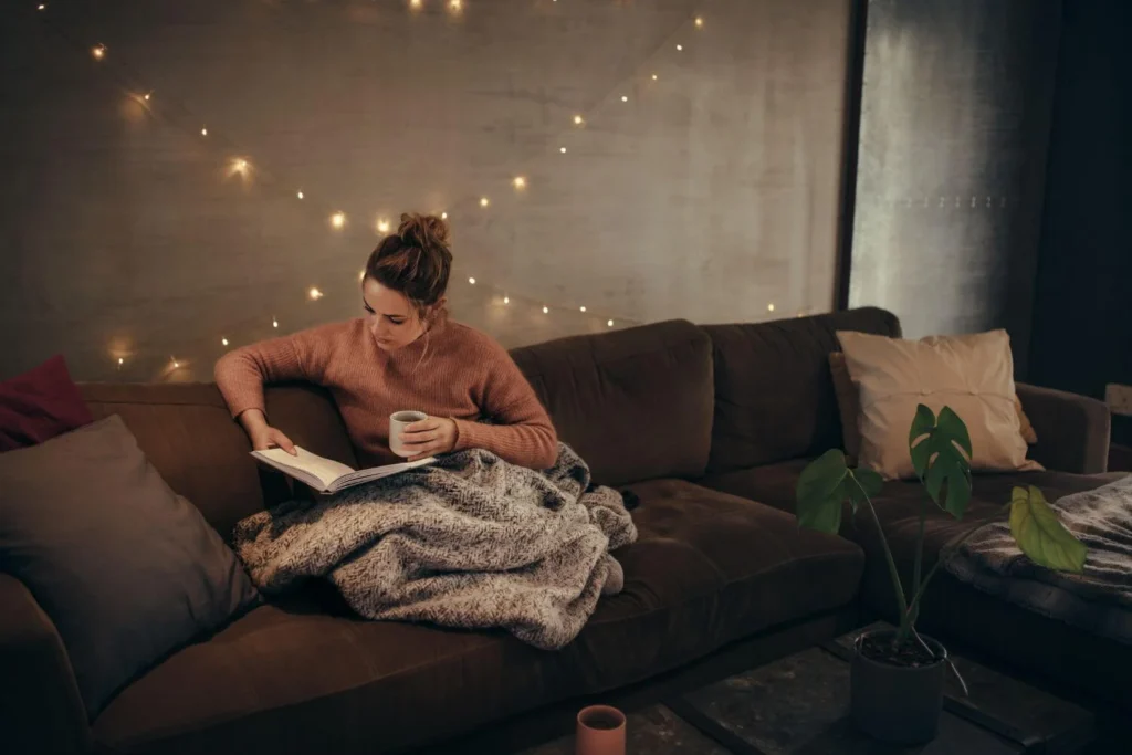 Woman relaxing on a sofa with a warm blanket, reading a book and holding a mug, with soft Christmas fairy lights glowing in the background.