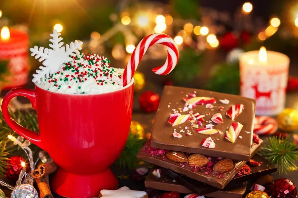 Bright red mug filled with whipped cream, holiday sprinkles, a candy cane, and a white snowflake topper next to festive chocolate bark with crushed peppermint and almonds, surrounded by Christmas lights and candles.