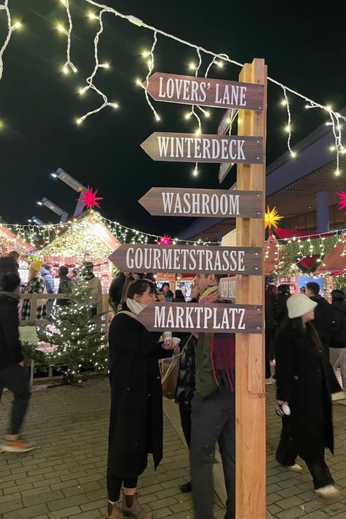 A wooden direction sign at a Christmas market pointing toward Lovers’ Lane, Winterdeck, and Marktplatz with twinkling lights overhead.