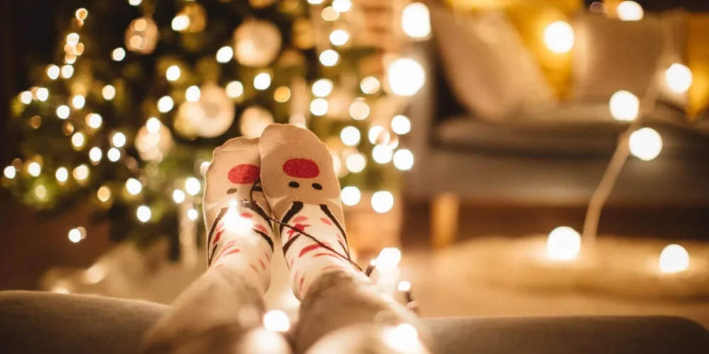 Person lounging in cozy reindeer socks with Christmas tree lights wrapped around their legs, blurred festive background showing a glowing Christmas tree and soft-lit living room.