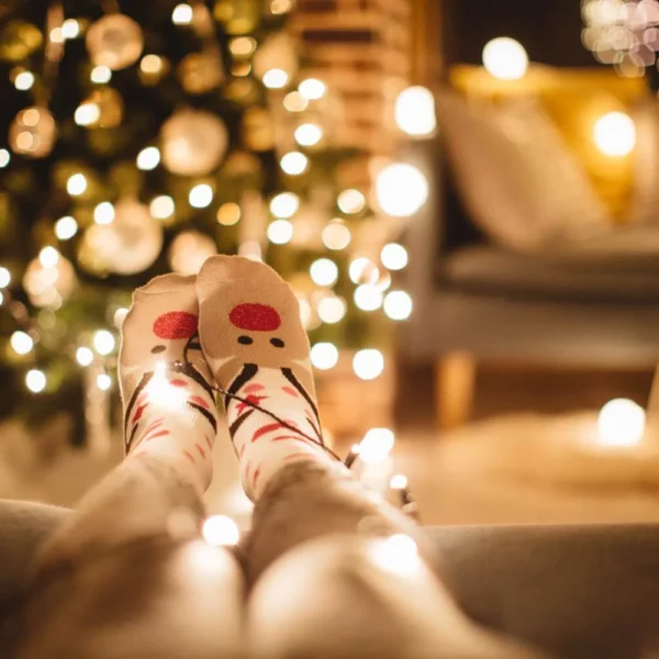 Person lounging in cozy reindeer socks with Christmas tree lights wrapped around their legs, blurred festive background showing a glowing Christmas tree and soft-lit living room.