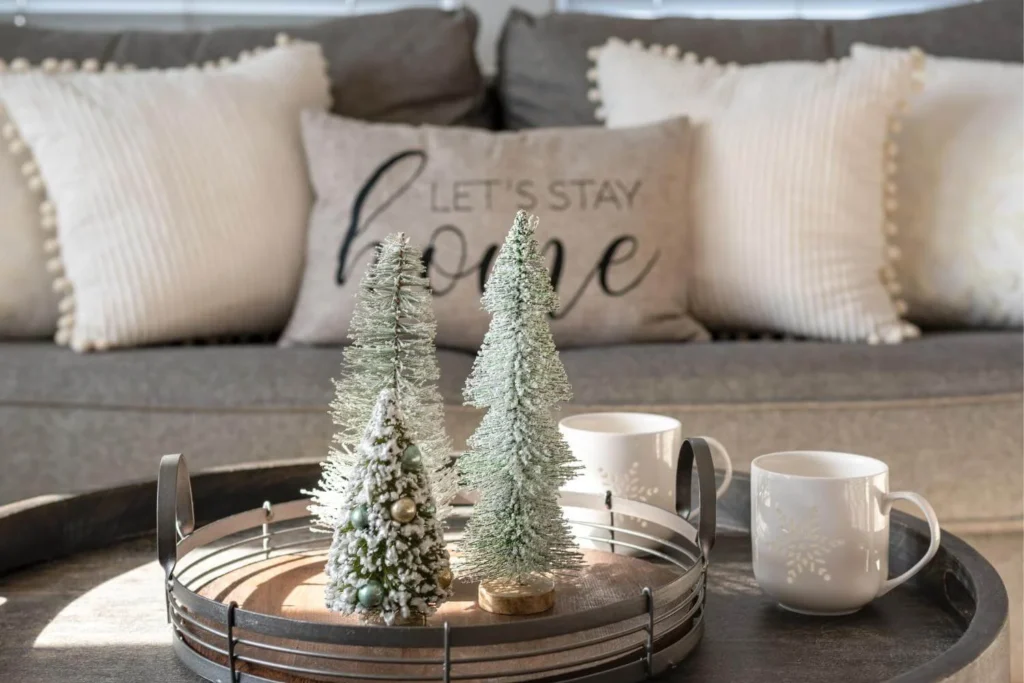 Small decorative Christmas trees and two winter mugs arranged on a tray in a cosy living room with soft cushions.