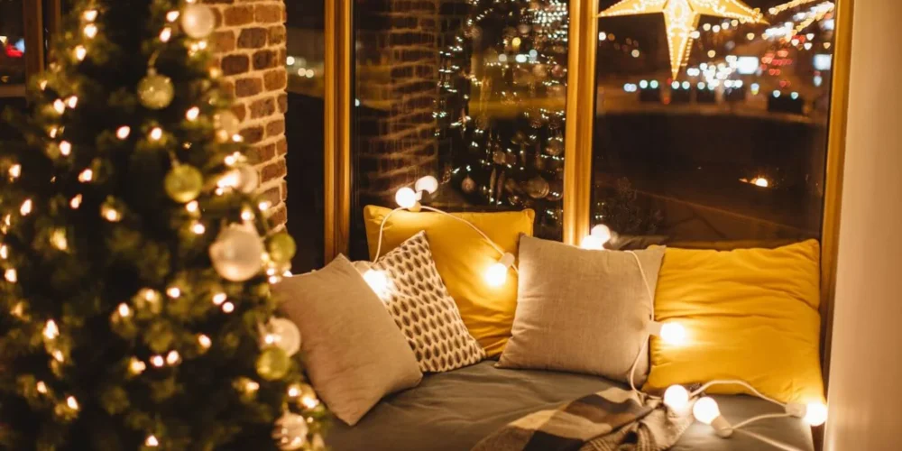 Warm and inviting window nook decorated for Christmas with soft cushions, fairy lights, a hanging star lantern, and a plaid blanket, next to a glowing Christmas tree reflected in the window.