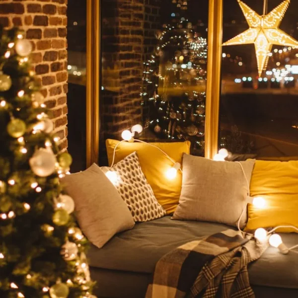 Warm and inviting window nook decorated for Christmas with soft cushions, fairy lights, a hanging star lantern, and a plaid blanket, next to a glowing Christmas tree reflected in the window.