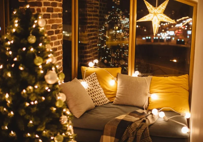 Warm and inviting window nook decorated for Christmas with soft cushions, fairy lights, a hanging star lantern, and a plaid blanket, next to a glowing Christmas tree reflected in the window.