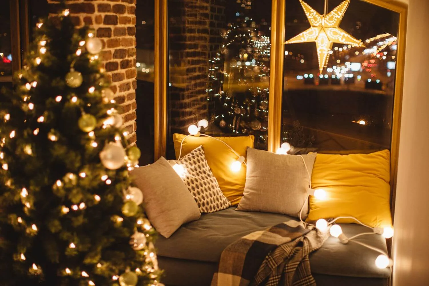 Warm and inviting window nook decorated for Christmas with soft cushions, fairy lights, a hanging star lantern, and a plaid blanket, next to a glowing Christmas tree reflected in the window.