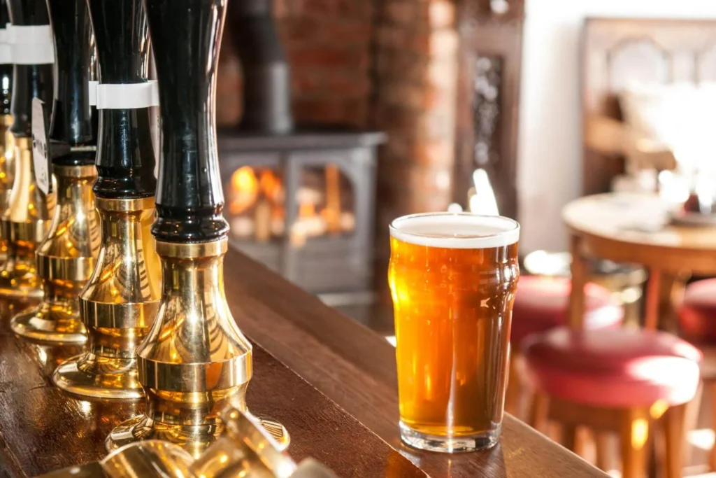 A pint of ale on the bar inside a cozy pub with brass taps and a glowing fireplace in the background.