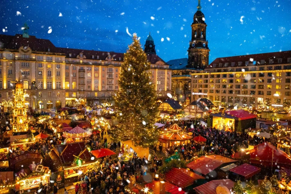 Bustling Christmas market at night in Dresden, Germany with glowing stalls, a large decorated tree, and light snowfall creating a festive winter atmosphere.