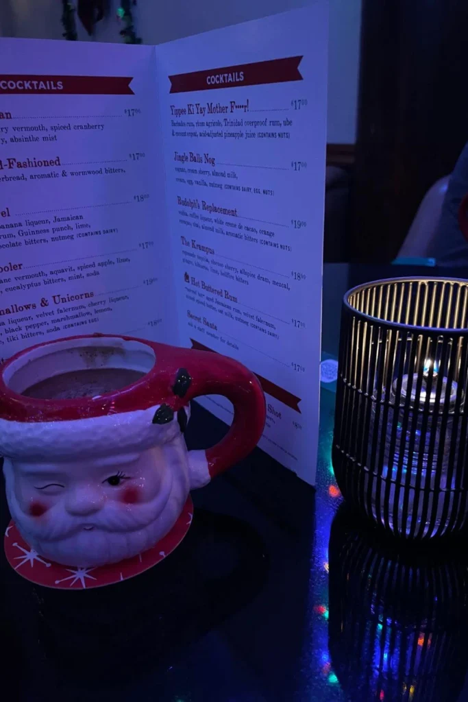 A cozy holiday cocktail served in a winking Santa mug on a candlelit table at a festive Christmas bar.
