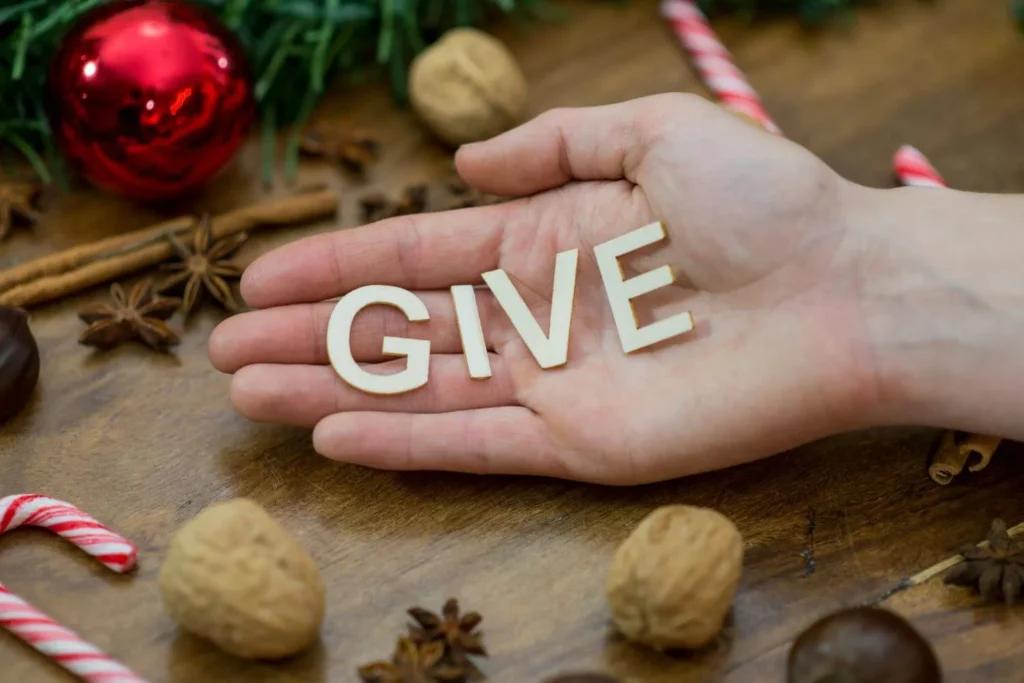 Hand holding wooden letters spelling GIVE surrounded by festive Christmas decorations and treats.