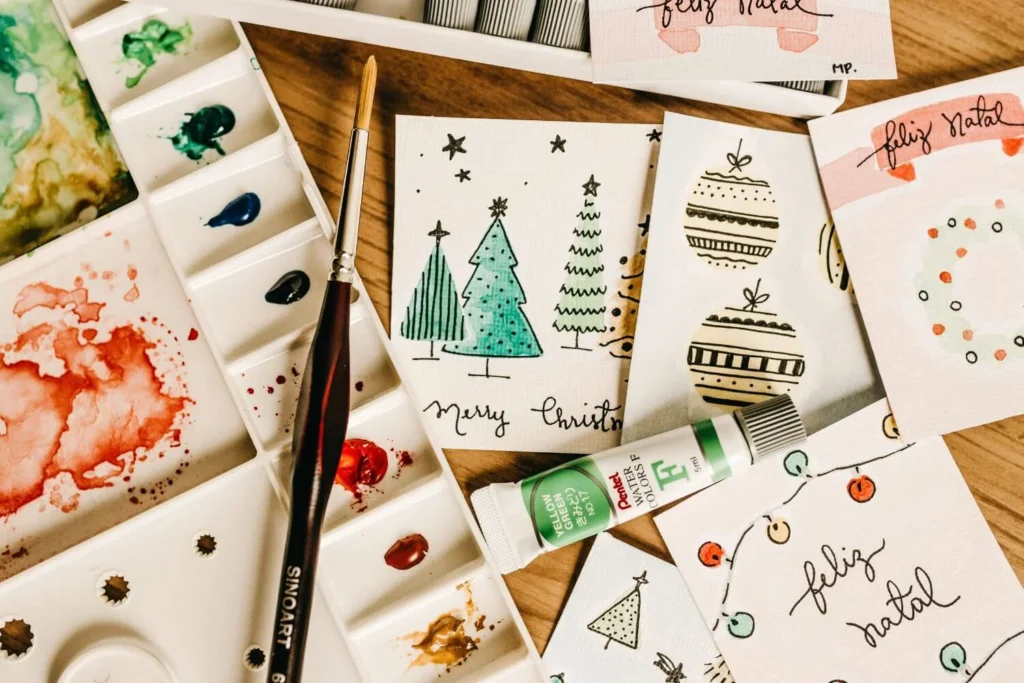 Flat lay of hand-painted Christmas cards with festive designs including trees, ornaments, wreaths, and fairy lights, surrounded by watercolor paints, a paintbrush, and a green paint tube on a wooden table.