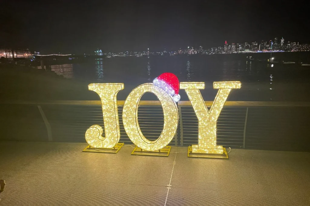 Illuminated JOY sign with a Santa hat glowing along the Vancouver waterfront at night.