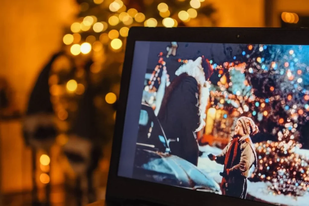 Laptop screen playing a Christmas movie with Santa and a child, set against a cozy background of warm fairy lights and a decorated Christmas tree.