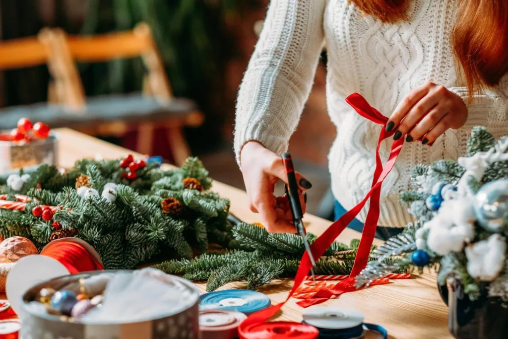 Woman in a white knit sweater cutting red ribbon while making a Christmas wreath with festive greenery and decorations.