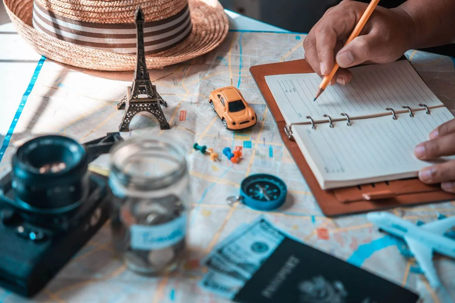 A travel journaling flatlay of a map, savings jar, money and a solo traveler writing in a notepad.