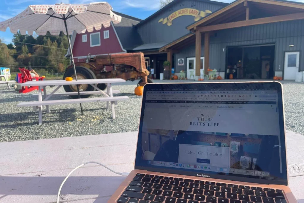 Laptop open to This Brits Life on an outdoor picnic table at Eagle Acres Dairy in Langley, with farm buildings, pumpkins, and a vintage tractor in the background, showing the idea of exploring your own city like a tourist.