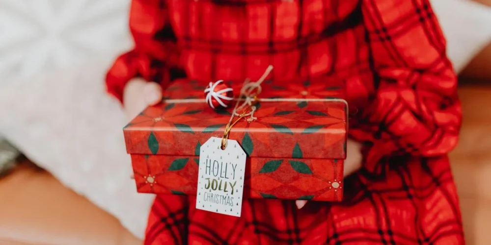 Woman in a red plaid dress holding a Christmas gift wrapped in red paper with a Holly Jolly Christmas tag.