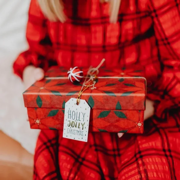 Woman in a red plaid dress holding a Christmas gift wrapped in red paper with a Holly Jolly Christmas tag.