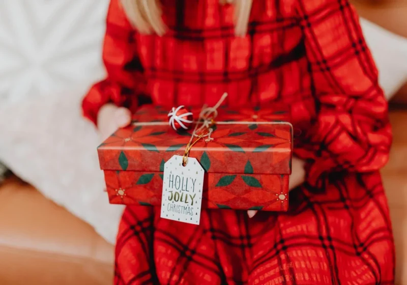 Woman in a red plaid dress holding a Christmas gift wrapped in red paper with a Holly Jolly Christmas tag.