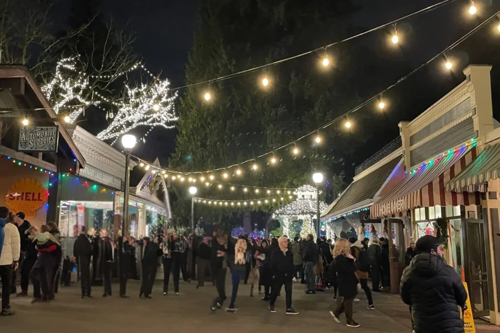 Festive heritage buildings at Burnaby Village Museum lit with colourful Christmas lights as visitors stroll through the outdoor village at night.