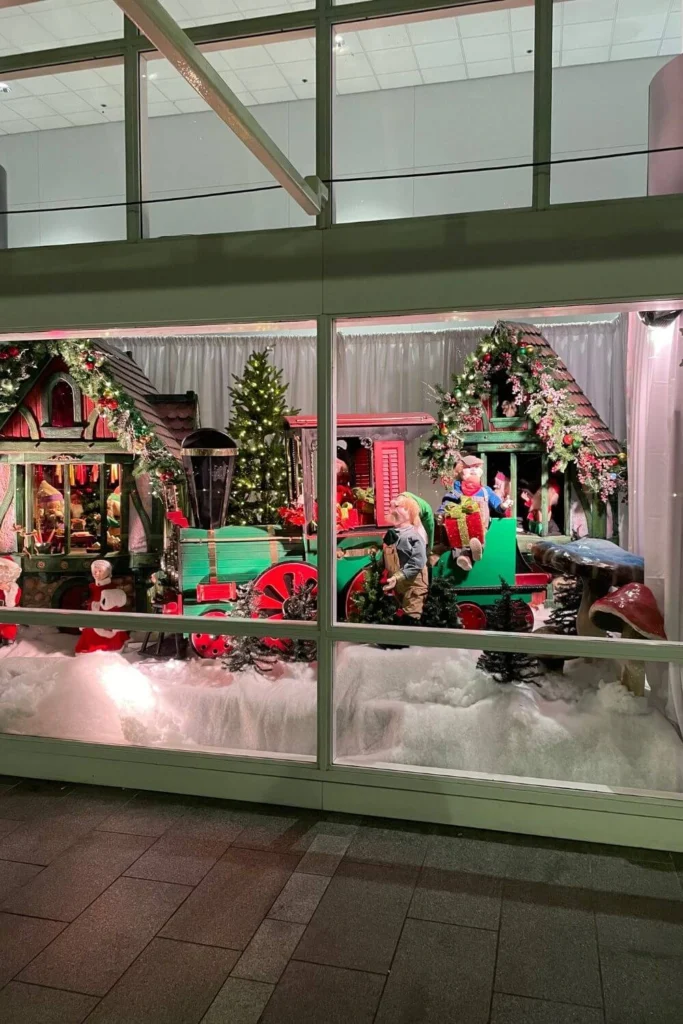 Whimsical Christmas window display at Canada Place with a green and red toy train, holiday figurines, and decorated village-style buildings surrounded by artificial snow.