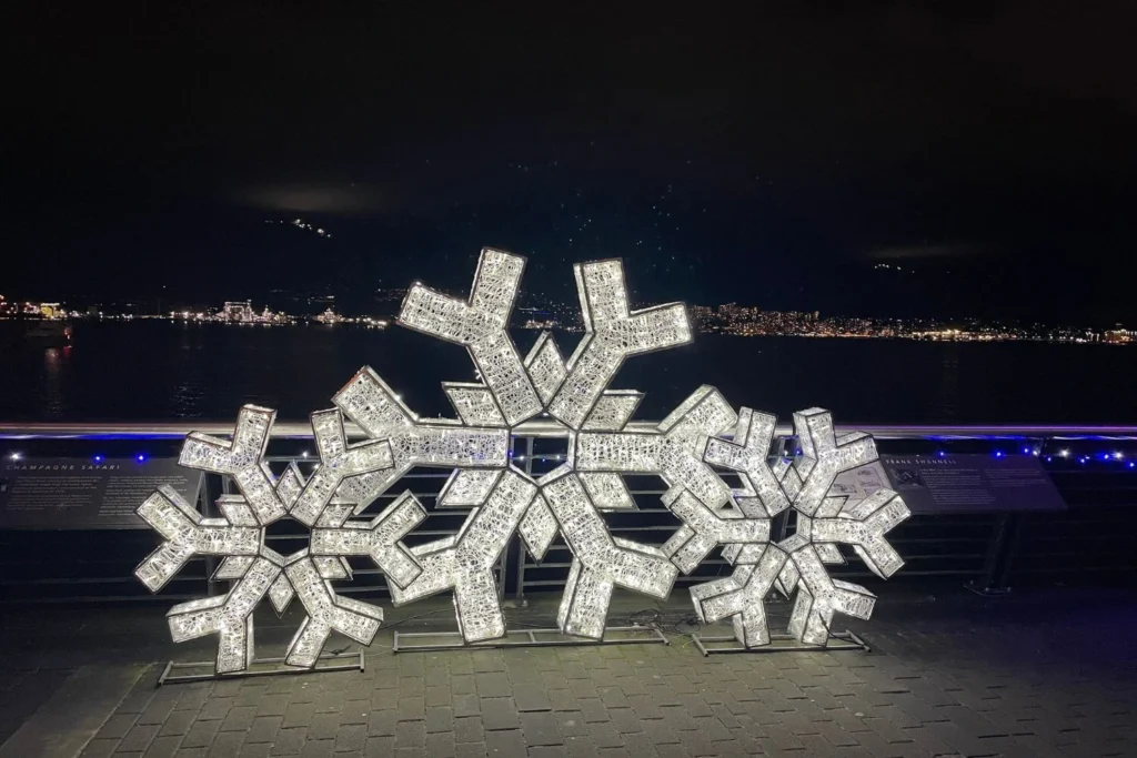 Cluster of three large snowflake light sculptures glowing in white along the Canada Place waterfront, with North Vancouver's lights twinkling across the harbor.
