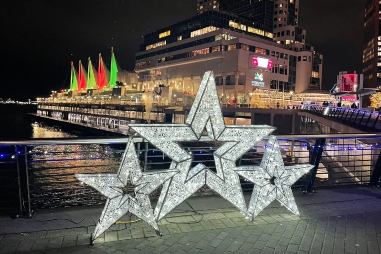 Three illuminated star sculptures on the Canada Place promenade, with sails of the convention center lit in red and green in the background for the holidays.