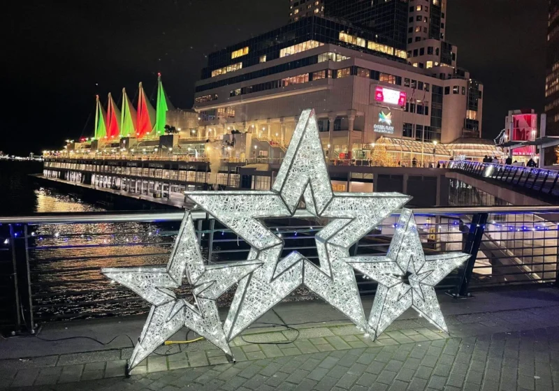 Three illuminated star sculptures on the Canada Place promenade, with sails of the convention center lit in red and green in the background for the holidays.