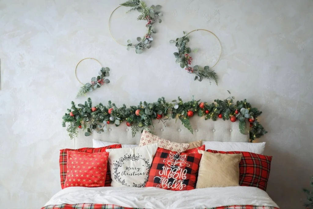 Festively decorated bed with red and plaid pillows, a garland of greenery and ornaments across the headboard, and minimalist wreaths on the wall above.